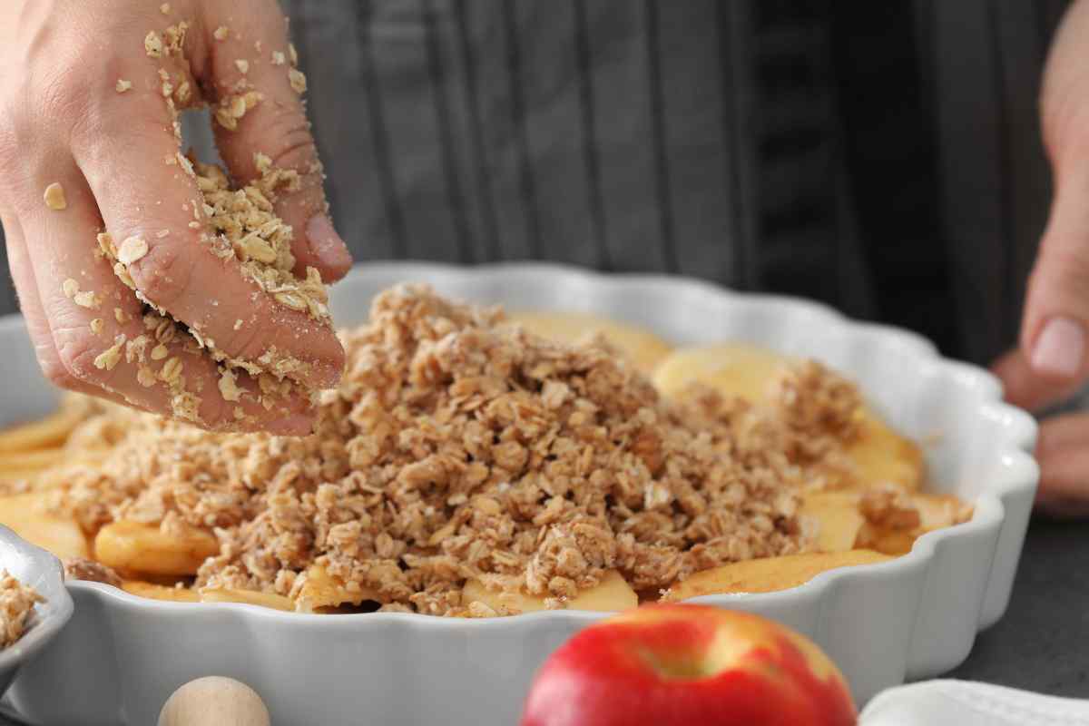 Image of hands adding a crumble mix to some softened apples in an oven dish , this is part of a blog about making apple crumble with kids