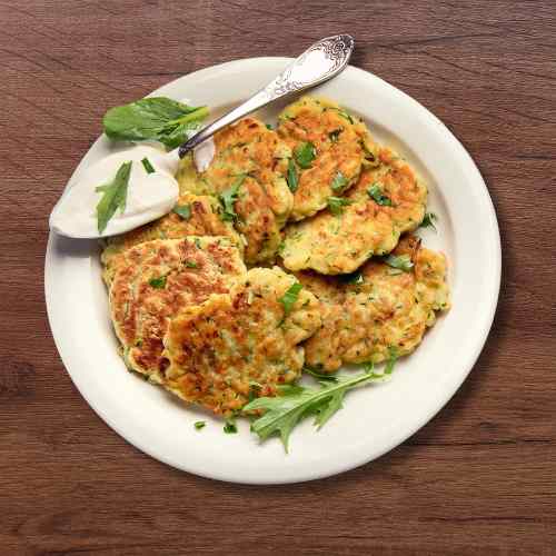 Image of some courgette cooked fritters on a white plate with a spoonful of yoghurt, as part of a blog about making cheesy courgette fritters
