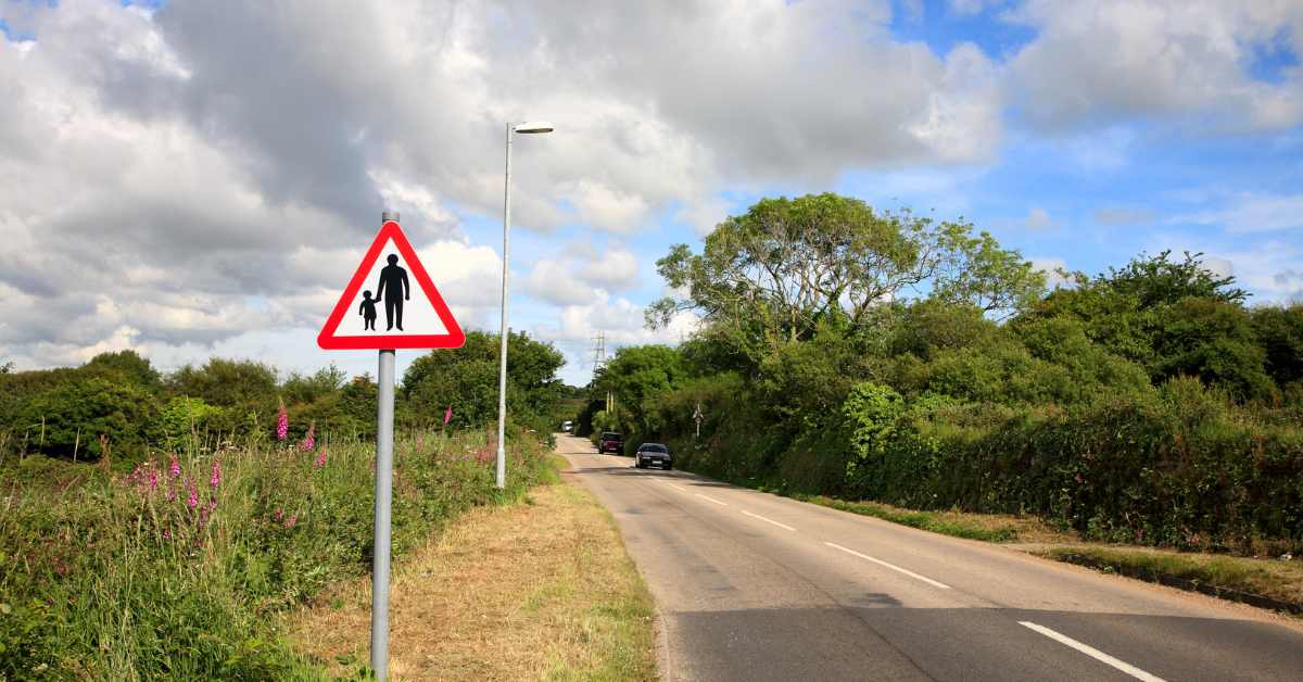 A country road with a couple of cars coming, but with wide verges and a hedges. A sign in the foreground of an adult and child to show that there is no pavement ahead. An image to help readers think about choosing a safe place to pick blackberries