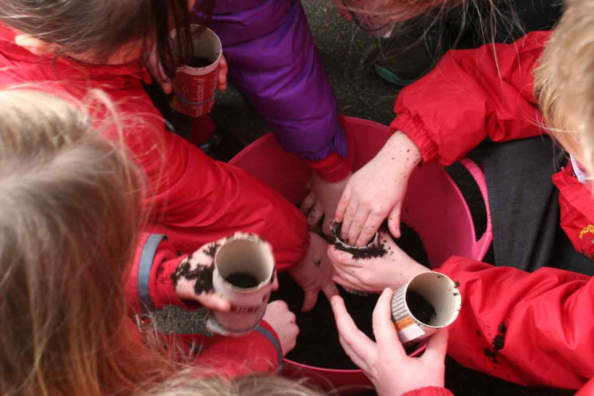 Image of children from overhead all leaning into a bucket of compost to gather handfuls of soil and push them into their toilet roll middles, this is part of a blog about sowing broad beans with kids