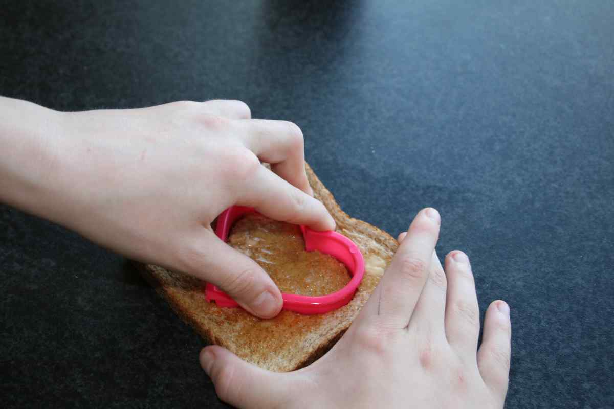 Picture of a child's hands cutting a hole out of some toast with a heart shaped cookie cutter, as part of a blog about a Valentine's recipe for kids to make independently called Love Toast.