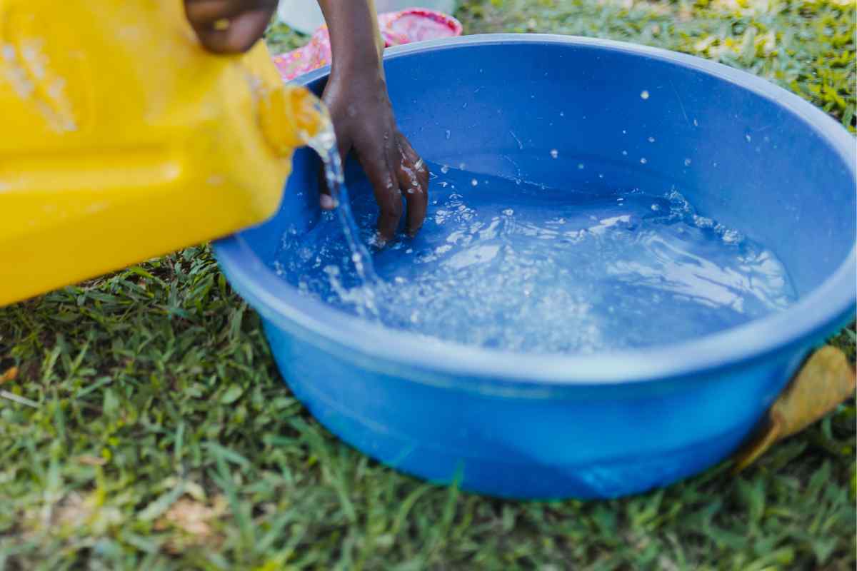 Image of a blue washing up bowl on some grass and someone pouring water from a canister into the bowl, this is part of a blog about washing plant pots with kids