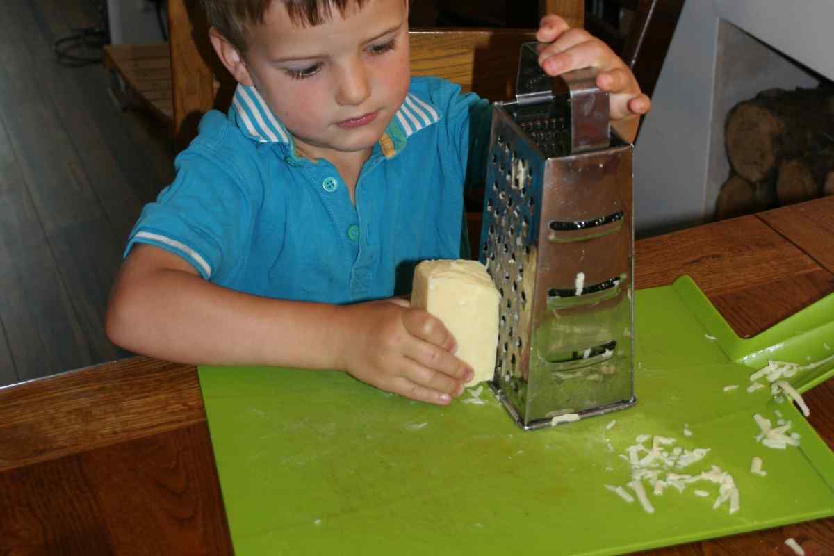 Image of a child carefully grating cheese with a box grater onto a board, this is part of a blog about making a leek and cheese pasta recipe with kids